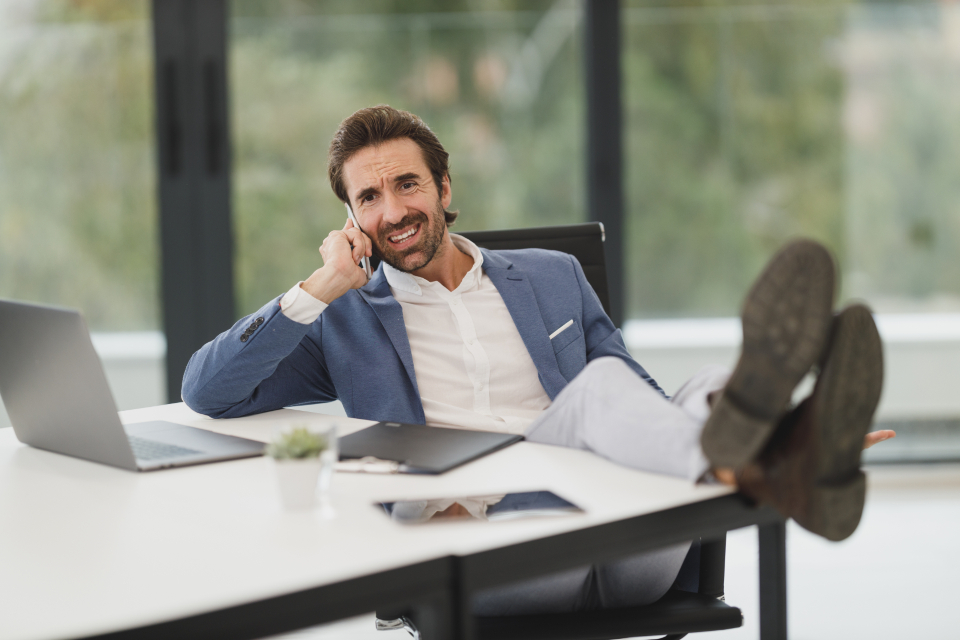 A man in a blazer on the phone with his feet on a desk