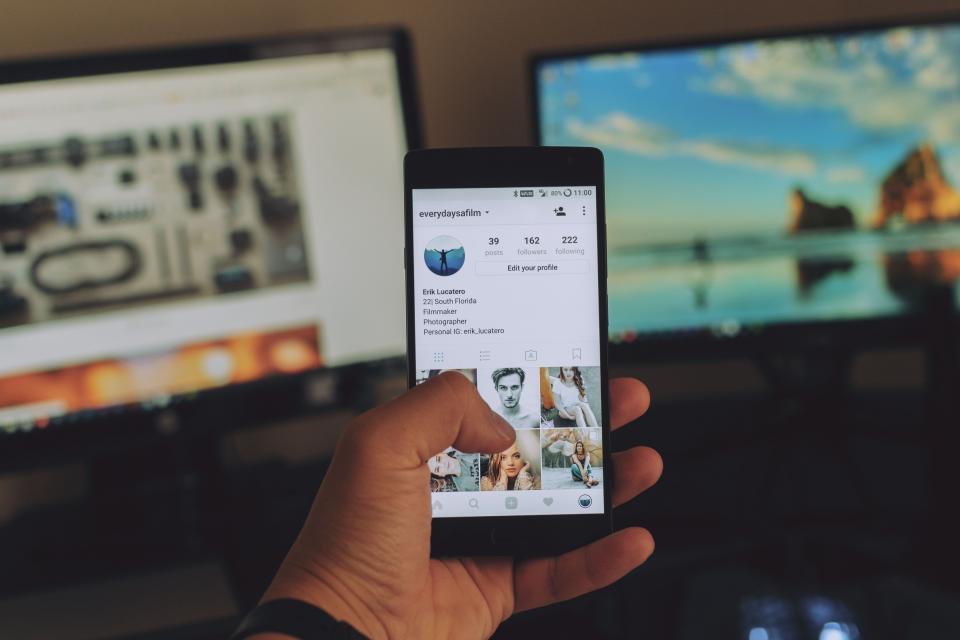 A phone displaying a social media profile with monitors blurred behind it